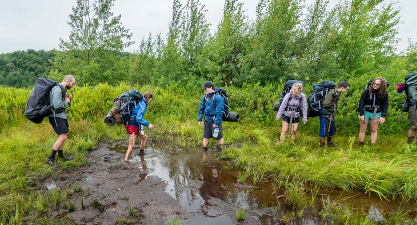 A group of young people wearing backpacks make their way though grass and ankle-deep mud.